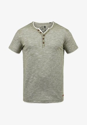 Lühikeste varrukatega, oliivroheline T-särk, mis on valmistatud tekstureeritud kangast. Omab valget kraed, kolme nupuga klappi ja väikest brändi silti alläärel.