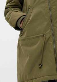 Parka vert olive avec une fermeture éclair à l'avant, des poignets élastiques et des poches latérales. Tissu lisse et mat avec un cordon de serrage à l'ourlet pour un ajustement.