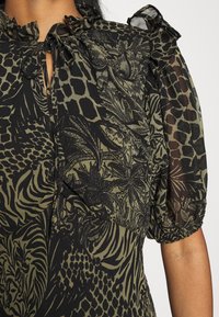 Svart och olivgrön blus med blommigt och djurmönster, med genomskinliga puffärmar och en nyckelhålsringning som är knuten med en rosett.