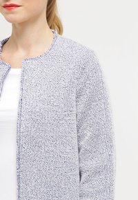 Texturerad ljusgrå cardigan i vävt tyg med rund halsringning och utan knappar. Har en enkel och stilren design.