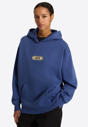 Młoda kobieta w niebieskiej bluzie Vans z logo na piersi i czarnych spodniach, stojąca z jedną ręką w kieszeni na białym tle.