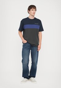 Menns kortermet T-skjorte i marineblå og grå, med en horisontal blå stripe og logo. Sammen med blå jeans og hvite sneakers.