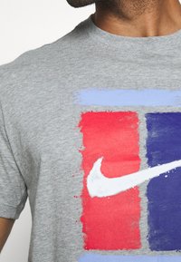 Graues T-Shirt mit einem grafischen Design aus roten, hellblauen und dunkelblauen Rechtecken; weißes Nike-Swoosh-Logo in der Mitte.