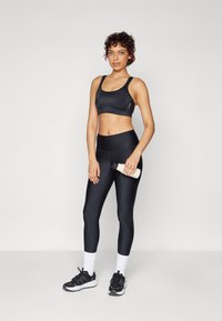 Svart sports-bh og høye tights laget av glatt stoff. Modellen holder en vannflaske. Hvite sokker og svarte joggesko fullfører antrekket.