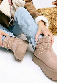 Stivaletti beige in suede, caratterizzati da un design pieghettato con dettagli borchiati. La suola spessa e texturizzata aggiunge altezza e stabilità.