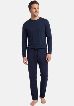 Mann steht barfuß, trägt ein marineblaues Langarmshirt und passende Hose mit Kordelzug vor weißem Hintergrund.