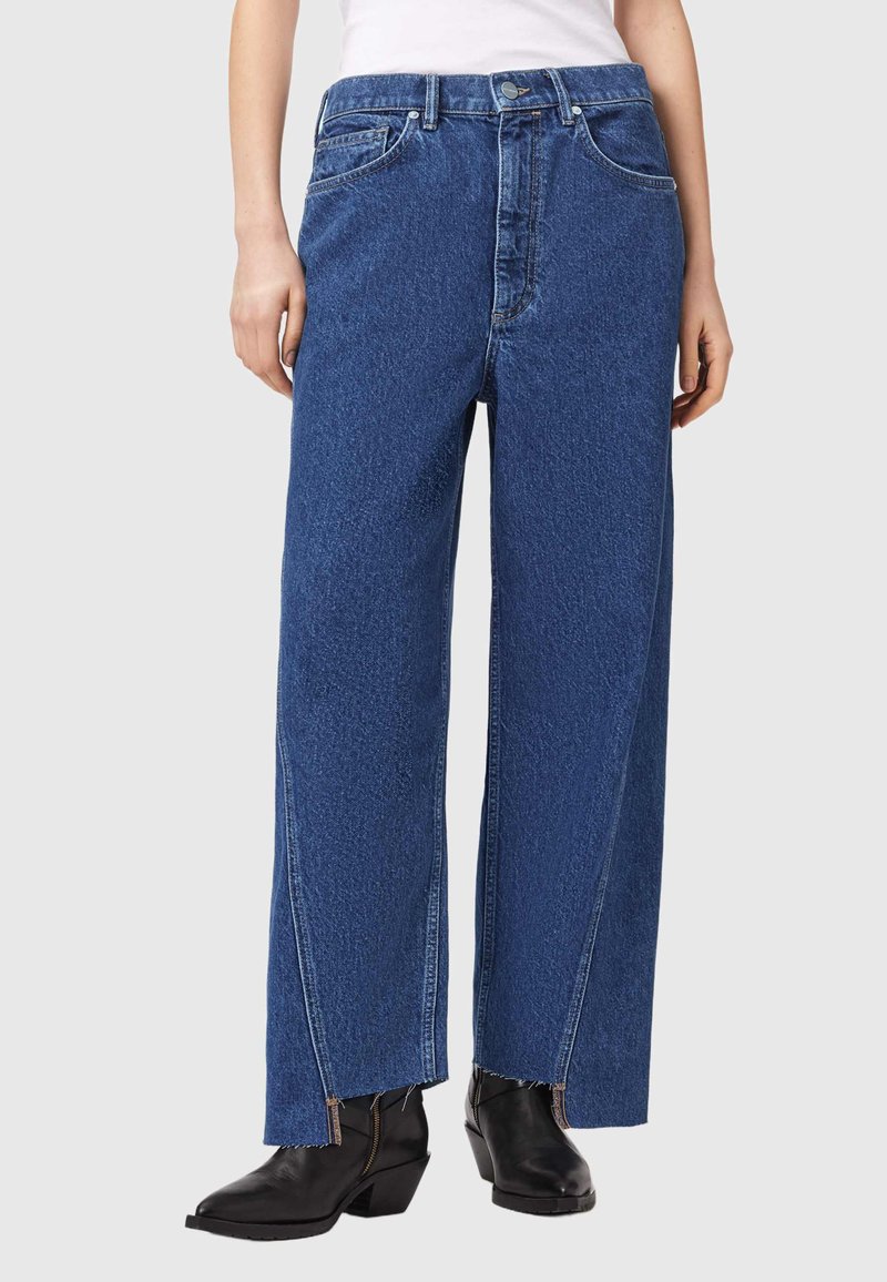 Persoon draagt blauwe denim jeans met wijde pijpen en frontale splitten en zwarte enkellaarzen, staand tegen een neutrale achtergrond.