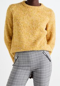 Persoon in een mosterdgele gestructureerde trui en hooggewaaid grijs geruite broek met zwarte ritszakken, staand voor een witte achtergrond.