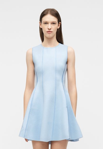 Femme aux longs cheveux bruns portant une robe bleu clair sans manches et ajustée avec des détails de coutures verticales, debout devant un fond uni.