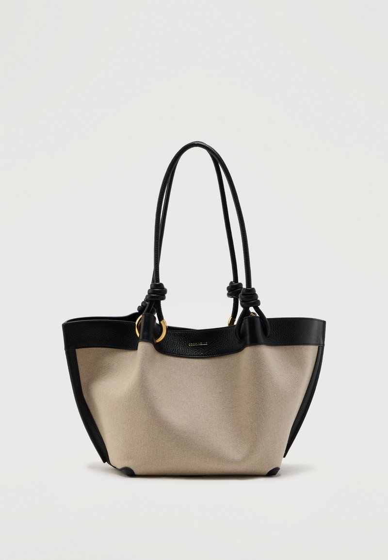 Μπεζ καμβάς τσάντα ώμου με μαύρη δερμάτινη επένδυση και διπλές λαβές, διακοσμημένη με χρυσές δακτυλίους και διακριτικό λογότυπο της επωνυμίας στο κέντρο πάνω μέρος.