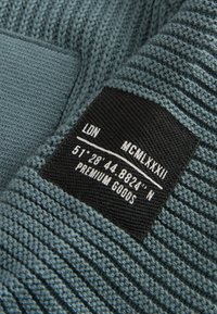 Étiquette en tissu noire sur un tissu tricoté bleu-gris avec un texte blanc affichant "LDN", des chiffres romains, des coordonnées et "Premium Goods".
