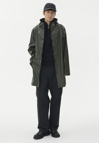 Grön vattentät jacka med huva, knäppning fram och en glänsande yta, ihop med svart ziptröja, svarta byxor och svarta snörskor.