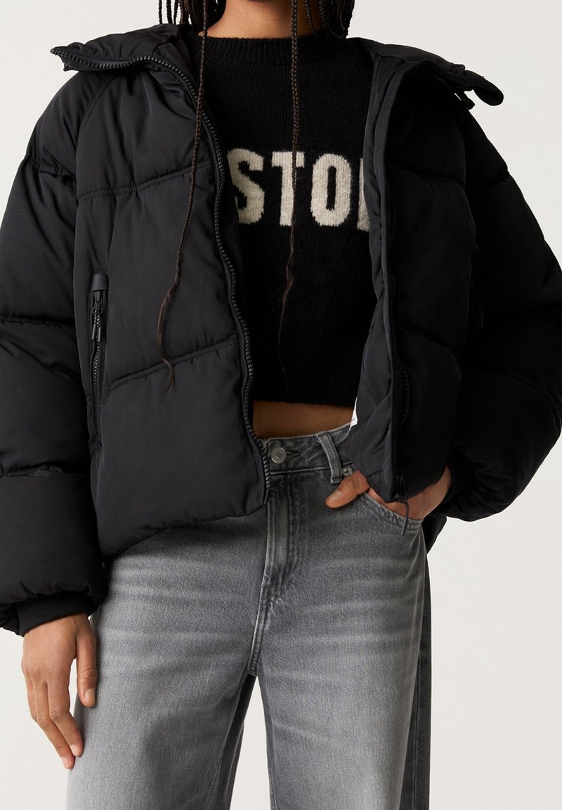 Sort pufferjakke med lynlås, der har hætte og kviltet design. Sammen med en sort sweater og grå jeans, der udstråler en afslappet stil.