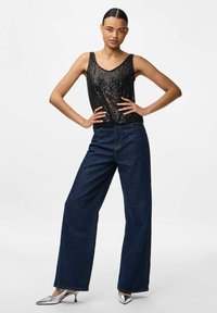 Giovane donna che indossa un top senza maniche nero con paillettes, jeans a gamba larga blu scuro, tacchi a punta argento, in posa con le mani sui fianchi.