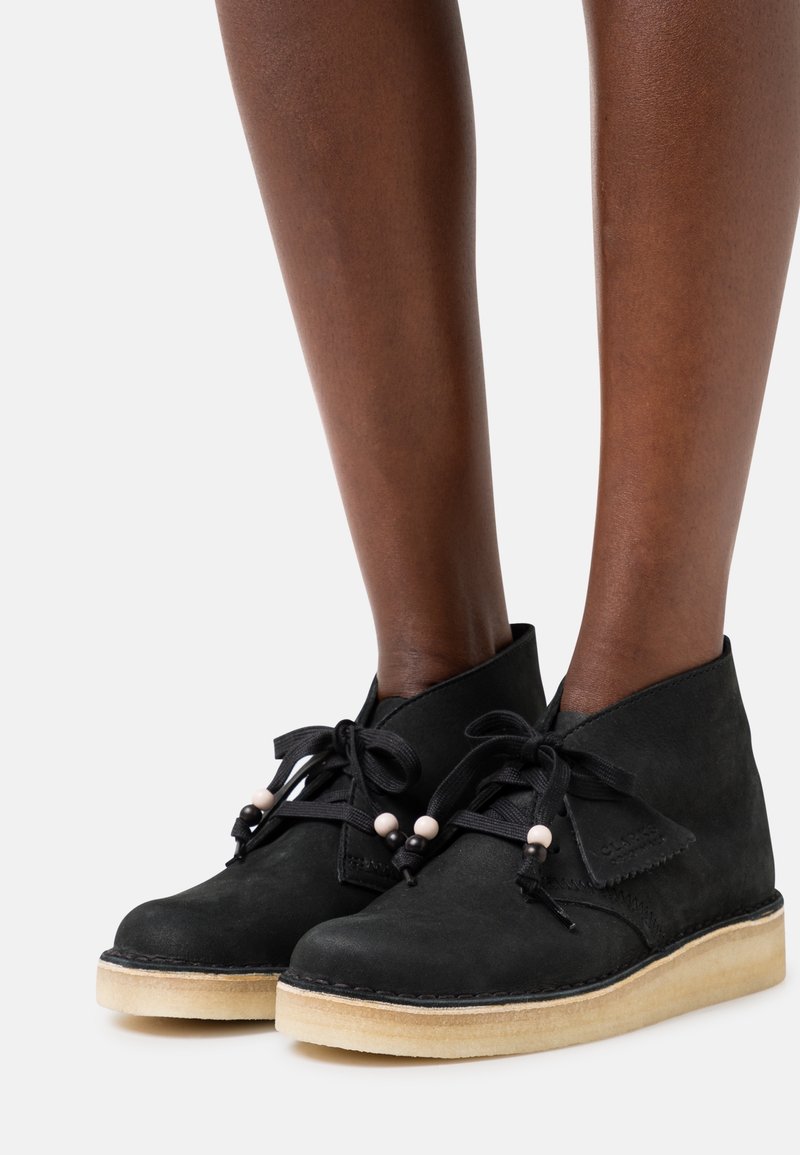 Bottes basses en daim noir avec un bout arrondi, dotées de lacets noirs avec des perles décoratives et une semelle en caoutchouc de couleur claire.