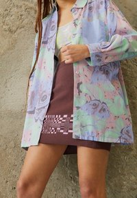 Mehrfarbiges, leichtes Hemd in Pastellfarben mit Blumen- und Schmetterlingsmotiven, kombiniert mit einem braunen Rock mit geometrischem Strickmuster.