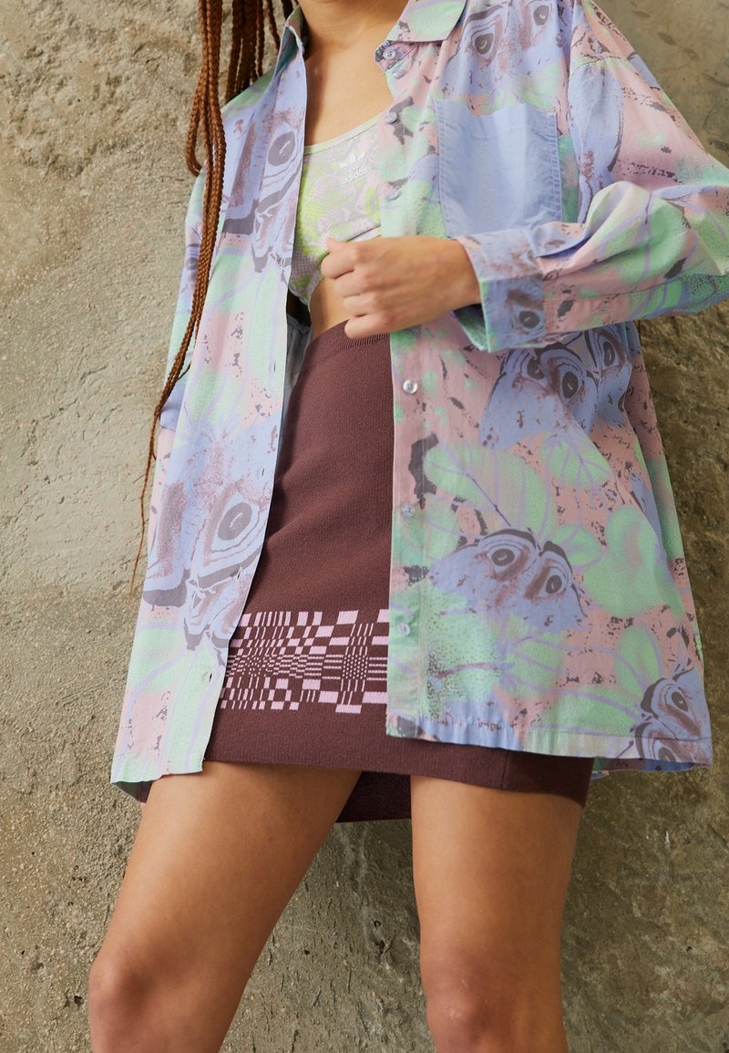 Mehrfarbiges, leichtes Hemd in Pastellfarben mit Blumen- und Schmetterlingsmotiven, kombiniert mit einem braunen Rock mit geometrischem Strickmuster.