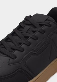 Μαύρο αθλητικό παπούτσι με επάνω μέρος από υφή δέρματος, μαύρα κορδόνια και ταμπά λαστιχένια σόλα, που δείχνει λεπτομερή ραφή και ανάγλυφη σήμανση στο πλάι.