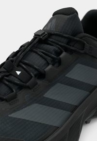 Černá atletická bota s texturovanou tkaninou, proužkovaným vzorem a tvarovanými detaily. Má rychlošněrovací systém a gumovou podrážku s dezénem.