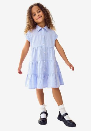Blauwe en witte geruite jurk met korte mouwen, gelaagd ontwerp en knopen aan de voorkant. Zwarte lakleren schoenen en witte sokken maken de outfit compleet.