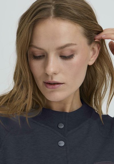 Tamsiai mėlynas marškinėliai su nuspaustu apykaklė, turintis priekinį kišenę ir du mygtukus. Audinys atrodo tekstūruotas, pabrėžiantis laisvalaikio dizainą.