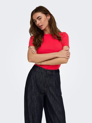 Femme aux longs cheveux bruns portant un haut court à manches courtes rouge vif et un jean large foncé, debout les bras croisés devant un fond uni.