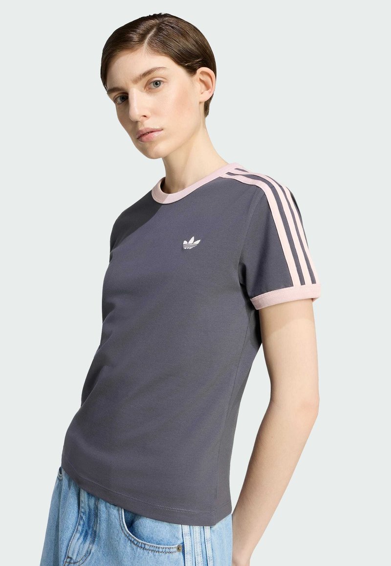 Kvinde iført en mørkegrå Adidas t-shirt med lyserød krave og striber, kombineret med lyseblå denimbukser, poserer mod en lys baggrund.