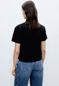 Black short-sleeve top with perforated texture and round neckline, paired with high-waisted light blue denim jeans.