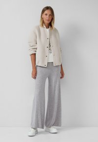 Beige gebreide jas met ronde hals en gouden knopen, gecombineerd met grijze wijde broekspijpen. Model draagt witte sneakers; neutrale achtergrond.