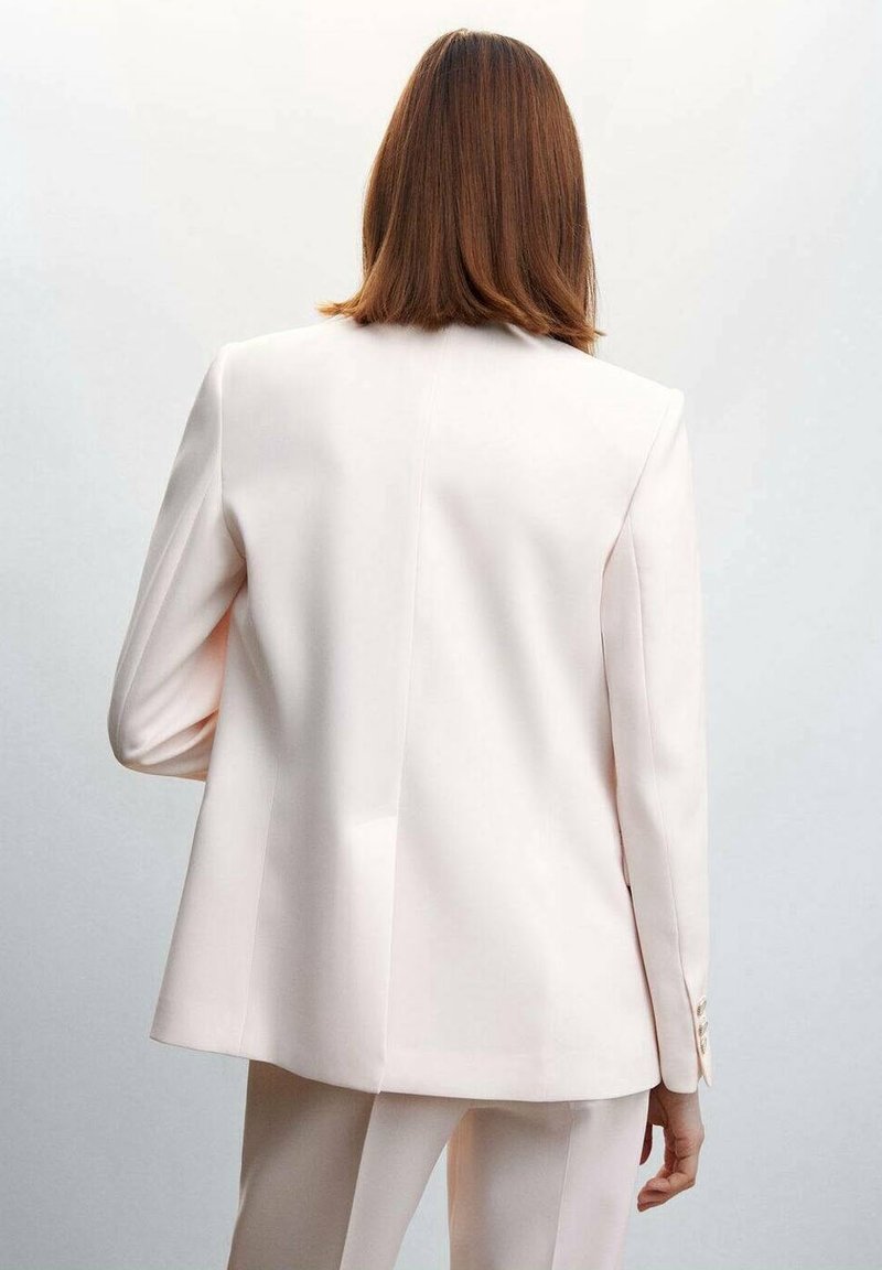 Femme aux cheveux bruns raides portant un blazer ajusté ivoire et un pantalon assorti, vue de dos sur un fond uni.