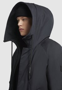 Chaqueta con capucha azul oscuro con un exterior texturizado, cordones ajustables y un cierre frontal con cremallera. Gorro de punto acanalado visible debajo de la capucha.