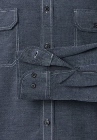 Blaues Hemd mit Knöpfen aus strukturiertem Stoff, mit dunklen Knöpfen, zwei Fronttaschen und kontrastierenden weißen Nahtdetails.