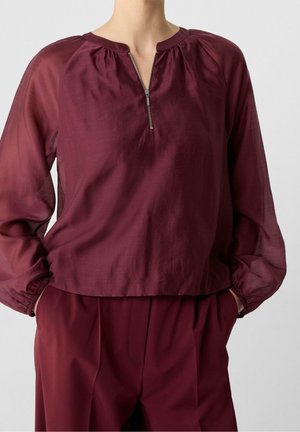 Femme portant un chemisier bordeaux à manches longues avec une fermeture éclair à l'avant et un pantalon taille haute assorti, mains dans les poches, sur un fond uni.