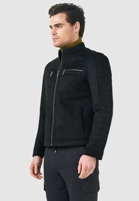 Giacca in suede nera con collo alto, dettagli trapuntati sulle spalle, due tasche sul petto con zip e chiusura con zip liscia.