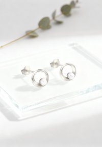 Silverfärgade, cirkulära stud earrings har en central vit pärla. Designen är minimalistisk med släta ytor och eleganta kanter.
