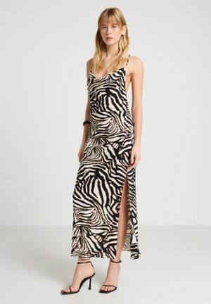 Donna che indossa un abito lungo senza maniche a stampa zebra con spacco laterale e sandali neri con tacco alto, in piedi davanti a uno sfondo neutro.