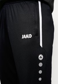 Zwarte sportieve broek met witte zijstrepen, voorzien van het zichtbare logo "JAKO" en ronde accenten bij de zoom voor extra detail.