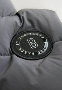 Tissu du manteau rembourré gris avec un badge noir rond arborant "Ted Baker" et une lettre "B" stylisée en blanc. Texture douce et matelassée.