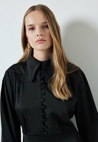 Black blouse with a collar and long sleeves, featuring a row of circular buttons down the front, made from a smooth fabric with a matte finish.