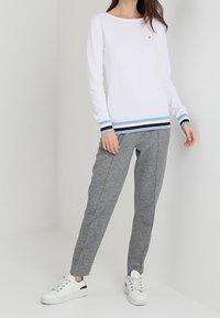 Wit overhemd met lange mouwen met blauwe en bruine gestreepte accenten aan de onderkant, gecombineerd met grijze joggingbroek en witte sneakers. Eenvoudig ontwerp.