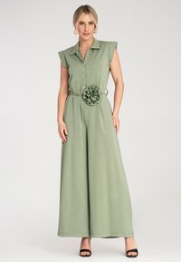 Zielony kombinezon bez rękawów z kołnierzykiem, guzikami z przodu, szerokimi nogawkami oraz tkaninowym kwiatem na talii. Gładka tekstura i casualowy design.