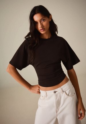 Femme aux longs cheveux foncés portant un haut noir moulant à manches courtes et un pantalon blanc taille haute sur un fond uni.