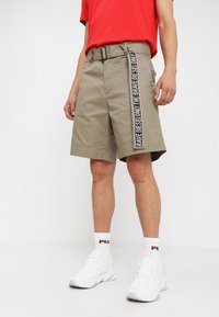 Shorts color khaki con una cintura e una fascia marchiata bianca e nera. Vita elasticizzata, davanti piatto, lunghezza fino a metà coscia. Abbinati a scarpe da ginnastica bianche.