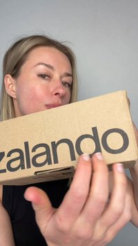 Vrouw met blond haar houdt een bruine Zalando kartonnen doos dicht bij haar gezicht tegen een effen lichtgrijze achtergrond.