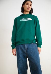 Hanorac verde cu buzunar frontal și logo alb Reebok, asortat cu blugi albastri, de tip lejer. Textură netedă, design casual.