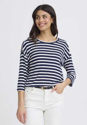 Frau lächelt, trägt ein marineblau-weiß gestreiftes Langarmshirt mit weißen Hosen und einem schwarzen geflochtenen Gürtel, eine Hand in der Tasche, schaut nach rechts.