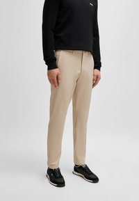 Pantaloni beige su misura con una texture liscia, vestibilità dritta, tasche laterali e dettagli di cucitura sottili, abbinati a sneakers nere.