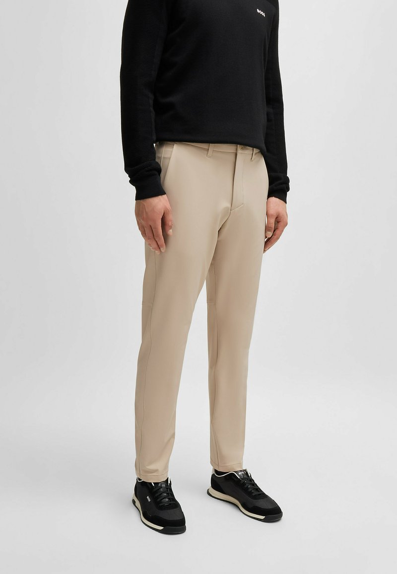 Pantaloni beige su misura con una texture liscia, vestibilità dritta, tasche laterali e dettagli di cucitura sottili, abbinati a sneakers nere.