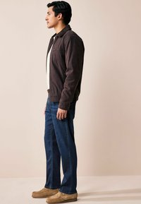 Jachetă din catifea maro, cu fermoar, tricou alb dedesubt, combinată cu blugi și pantofi slip-on bej deschis, văzută din lateral.