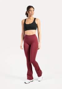 Femme en brassière de sport noire, leggings bordeaux taille haute, et baskets blanches, debout en appui sur une jambe, regardant vers la droite.